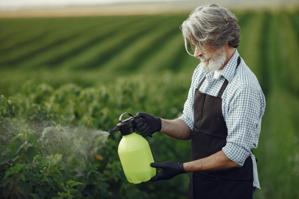 pexels-photo-4975361-4975361 Senior man in apron using spray bottle on lush green farmland during daytime, enhancing crop health.